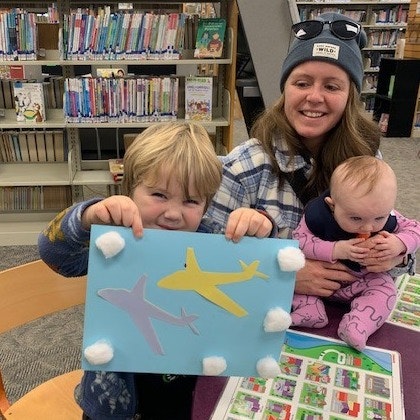 A child proudly displays a colorful art project featuring airplane cutouts, while a woman holds an infant in a library.