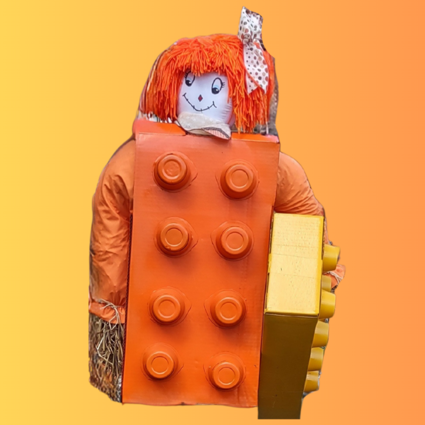 A scarecrow with a painted face and orange yarn hair, dressed in a costume resembling a large orange toy block.