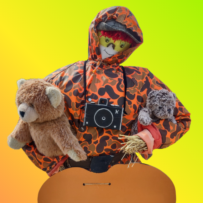 A scarecrow in a camo jacket with a camera and sunglasses, holding two stuffed animals, set against a vibrant green-yellow background.