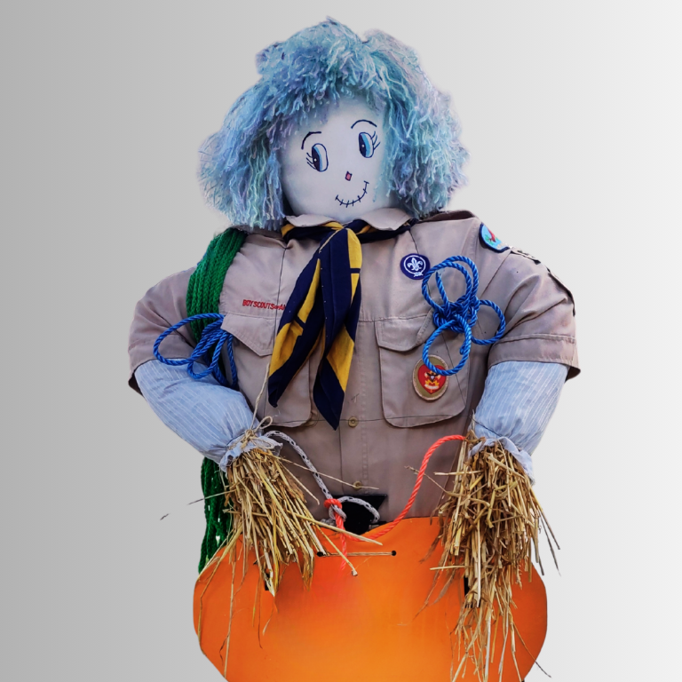 A scarecrow dressed in a scout uniform with a smiling face, blue yarn hair, and holding an orange object with straw hands.