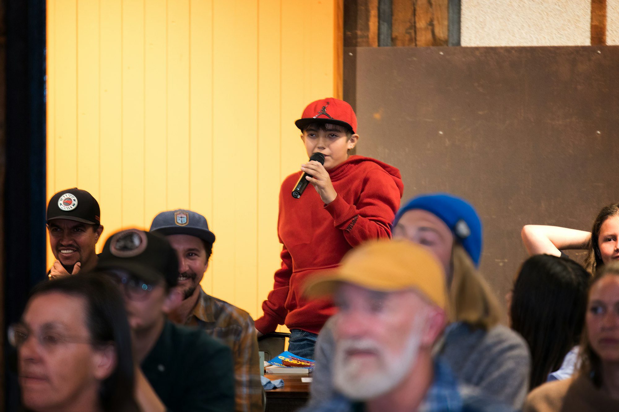 A person in a red hoodie and cap speaks into a microphone at an indoor gathering, surrounded by an attentive crowd.