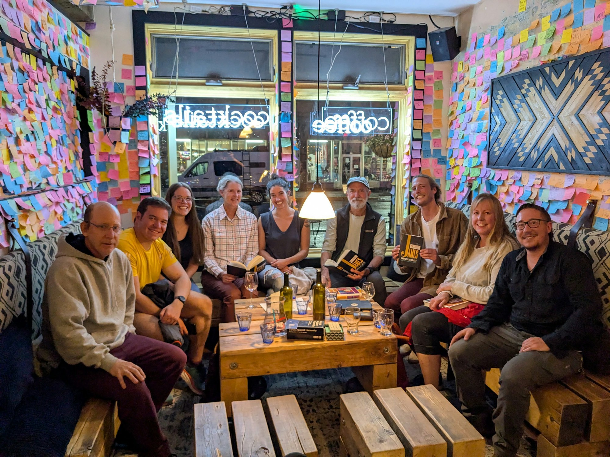 A group of eight people gathers in a cozy space with a wall covered in colorful sticky notes, sharing books and drinks.