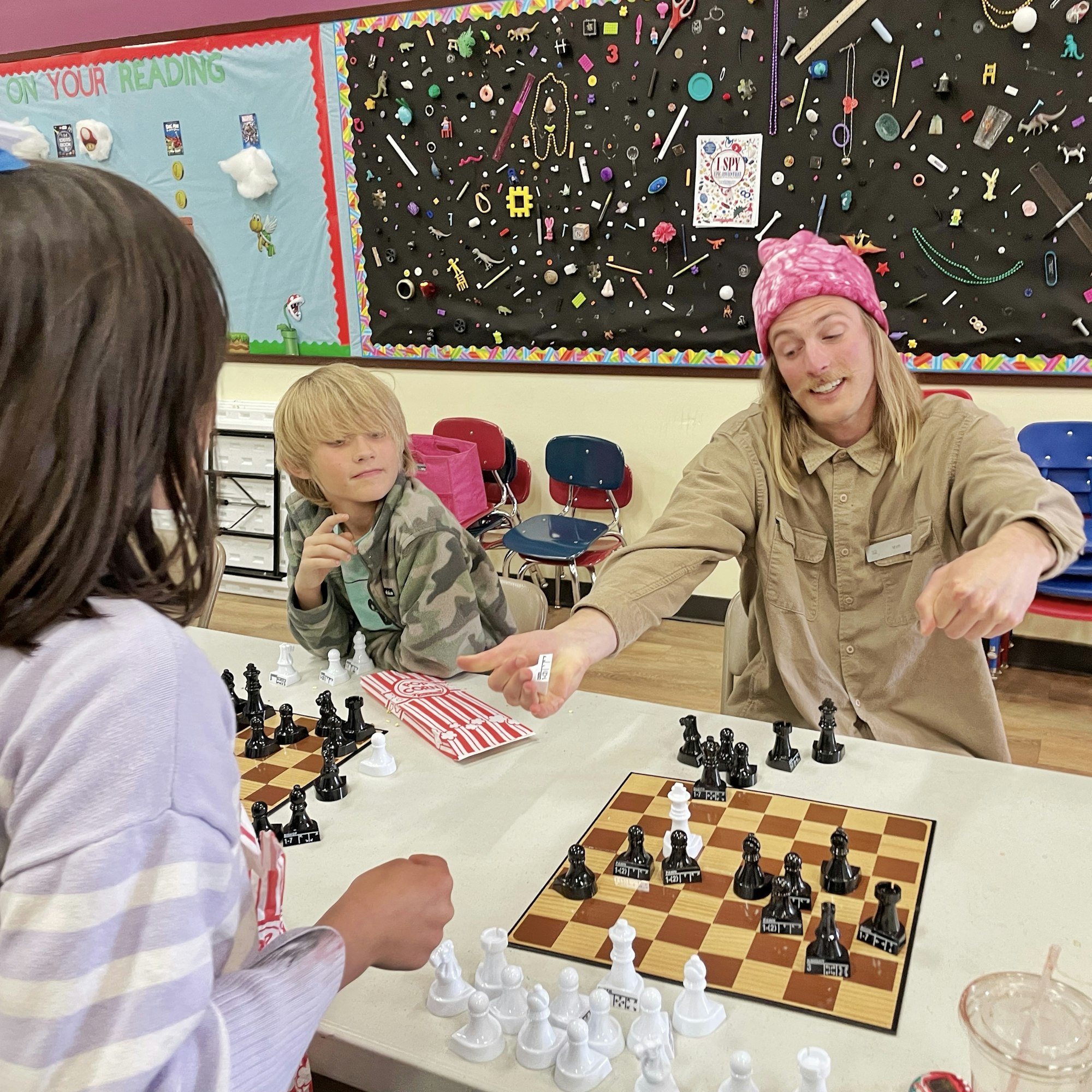 Three kids are playing chess at a table, with one teaching or explaining as they focus on the game. A colorful wall decor is in the background.