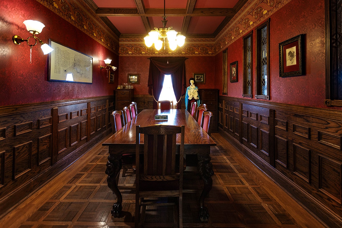 Victorian-style room with a long table, ornate wooden chairs, wall sconces, and a standing figure by a window.