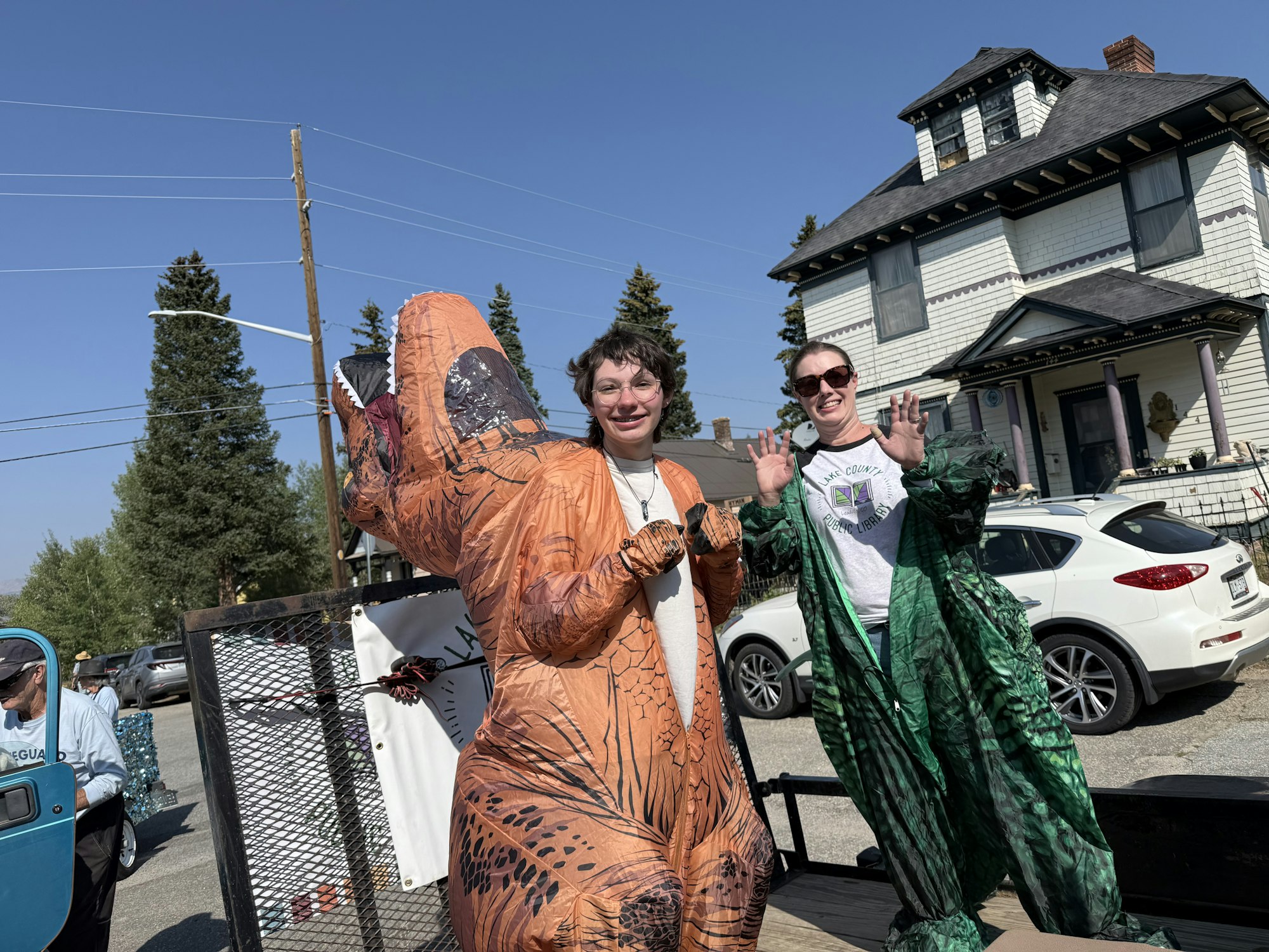 People in dinosaur costumes are having fun outdoors near a house, with a vehicle and some greenery in the background.