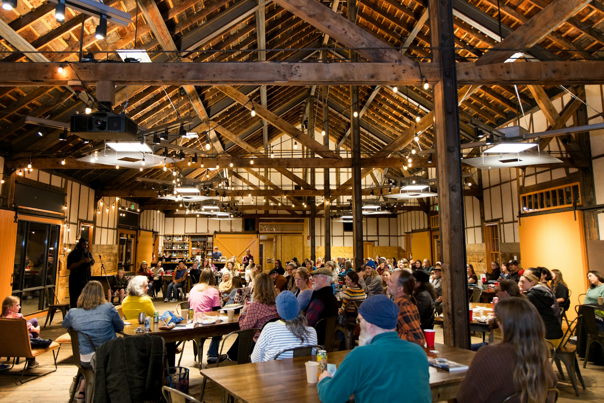A large group of people seated in a rustic, wood-paneled venue, listening to a speaker.