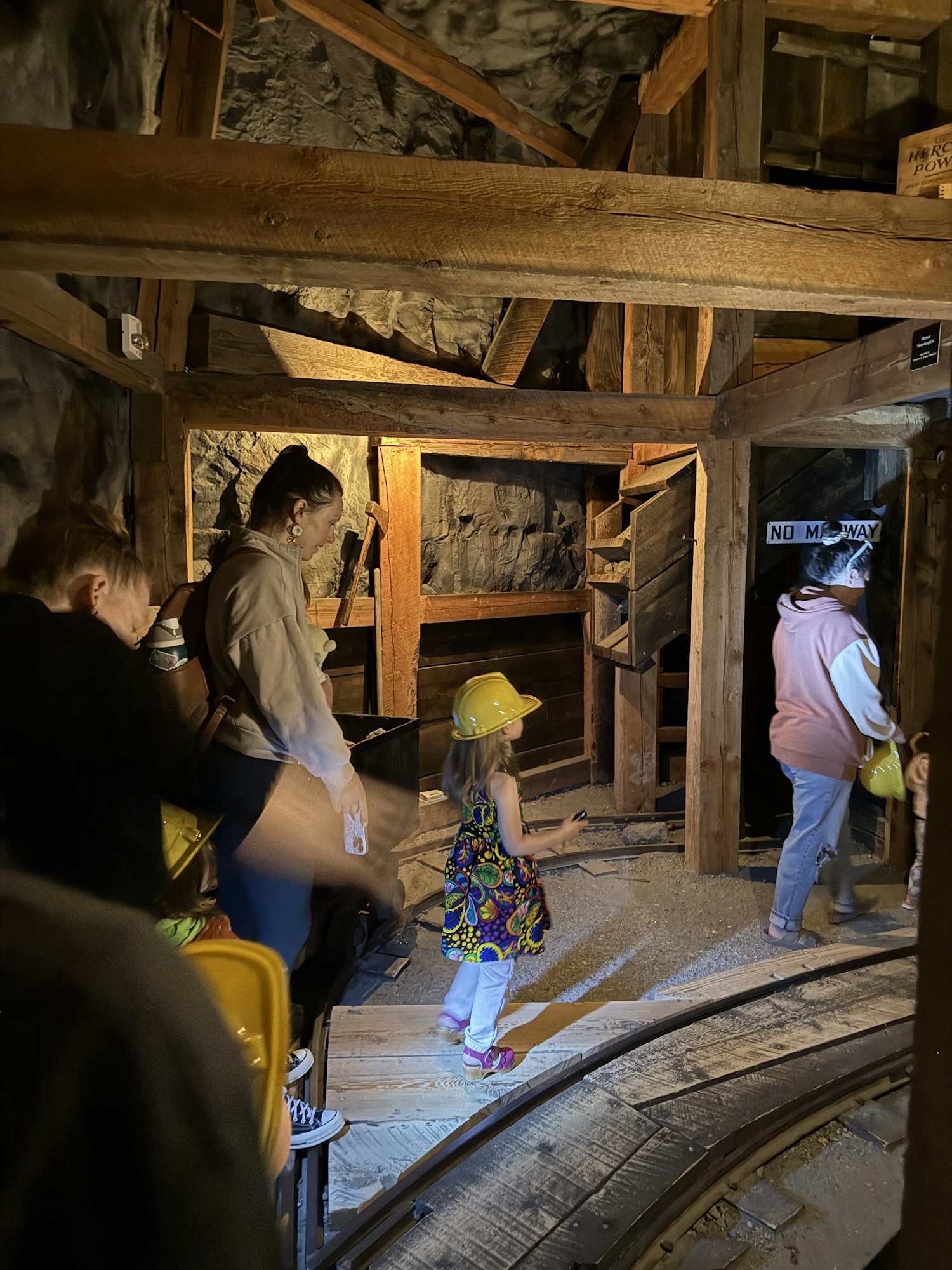 Inside a mine tour, people explore wooden structures, with a child in a hard hat and colorful outfit, while signs display warnings.