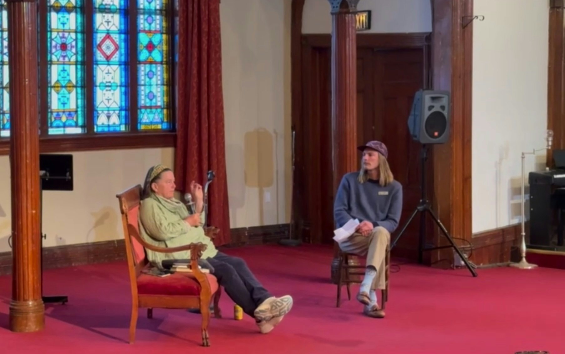 A seated conversation in a room with stained glass windows, featuring two individuals, one speaking and holding a microphone.