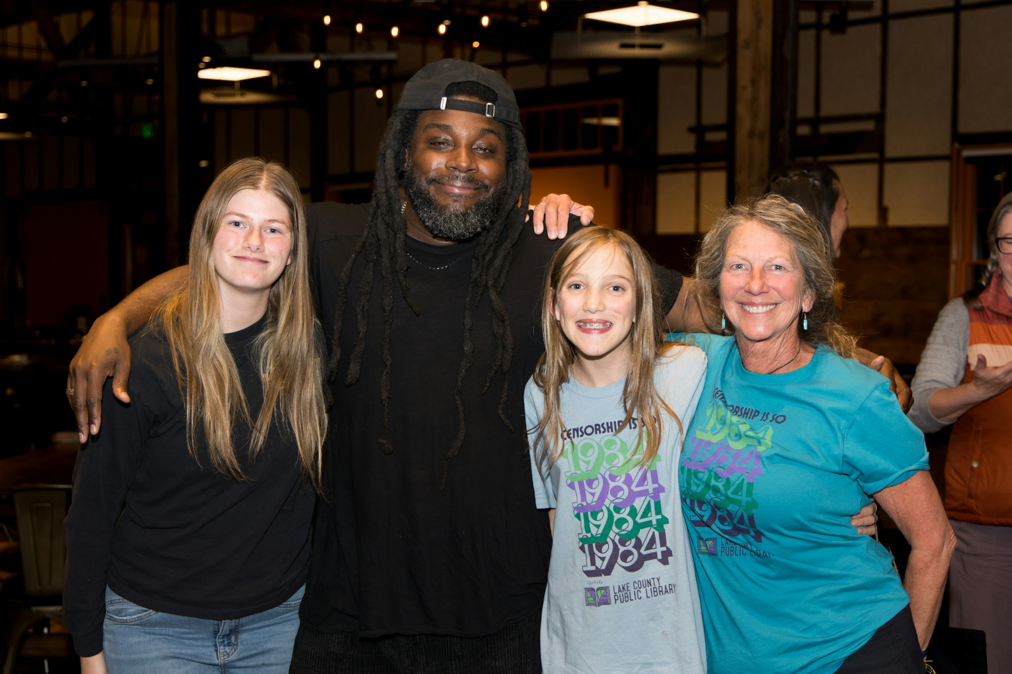 Four people smiling, standing together indoors; two wearing "Lake County Public Library" shirts.