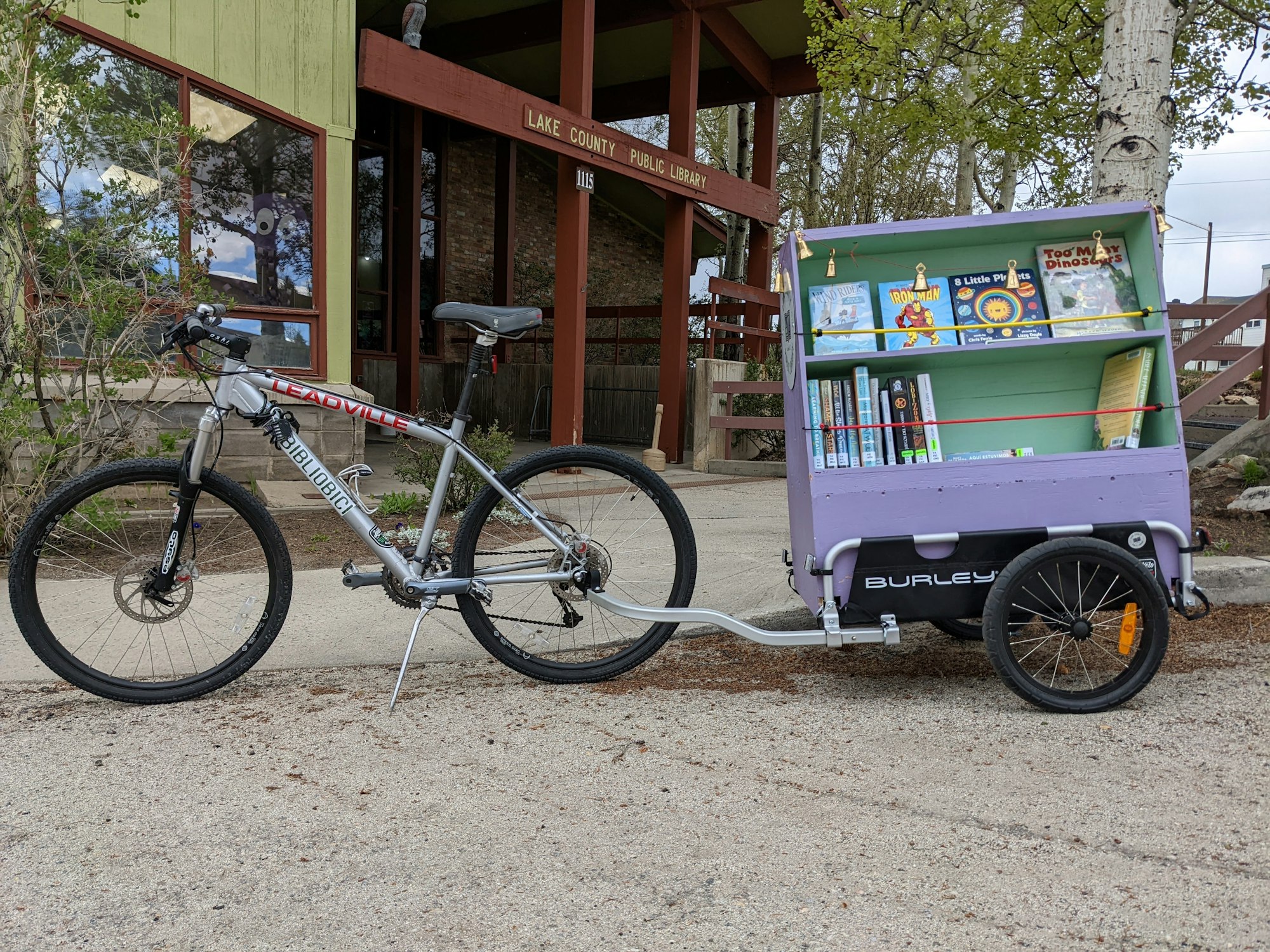 A bike with a trailer carrying books, parked outside a library.