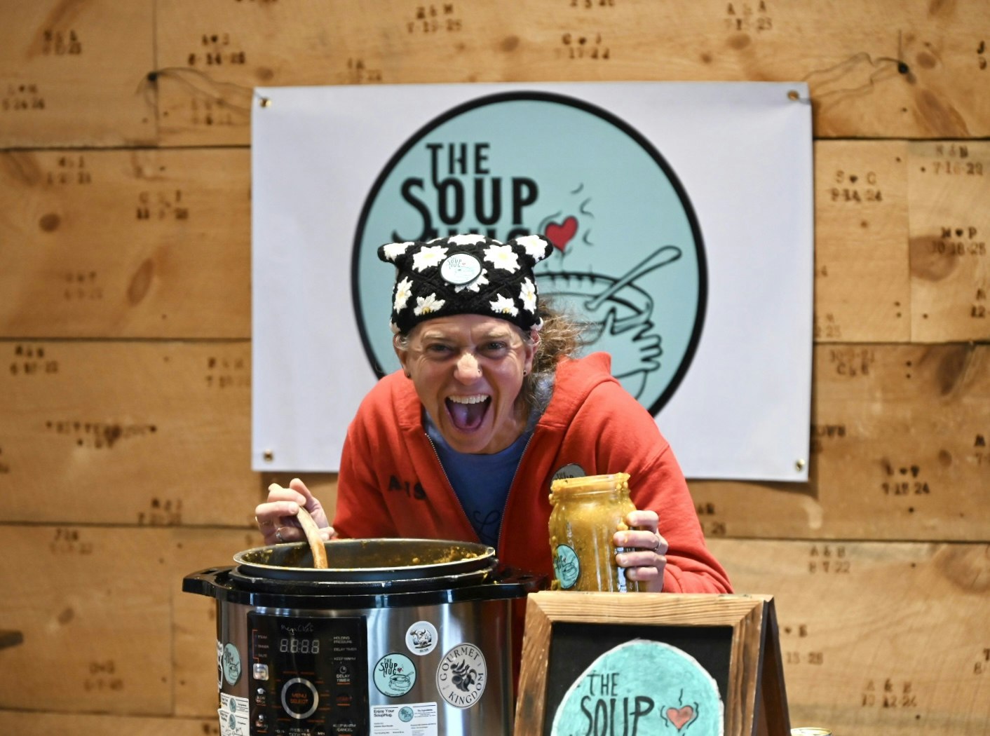 A cheerful person in a red hoodie is stirring a pot and holding a jar, promoting a soup-related theme.
