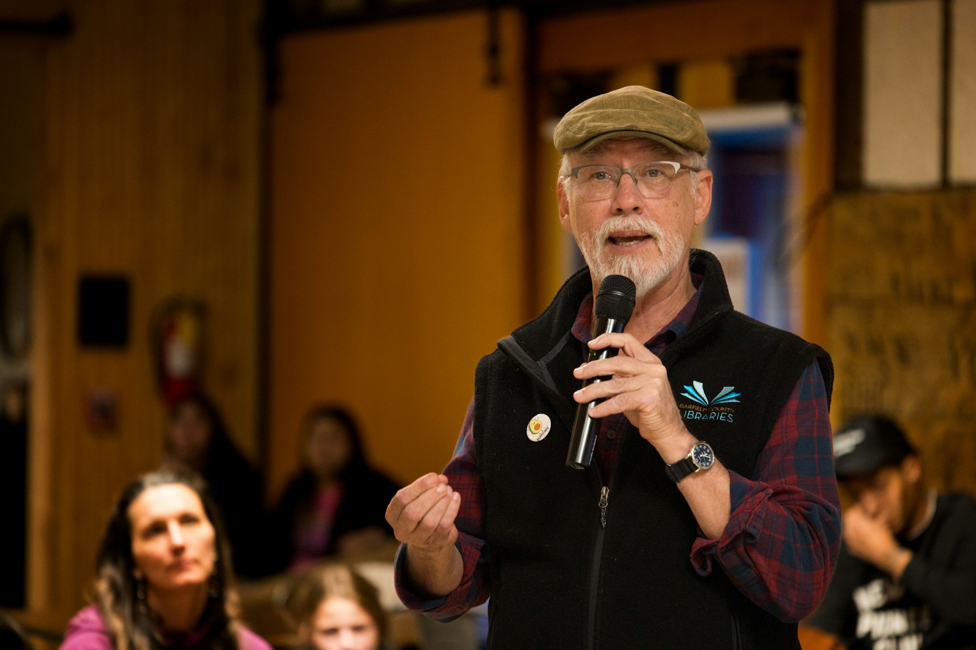 A man in a cap speaks into a microphone at an indoor event.