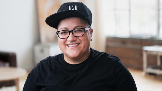 A smiling person (author Gabby Rivera) wearing glasses and a cap, sitting in a bright room with a casual outfit and friendly demeanor.