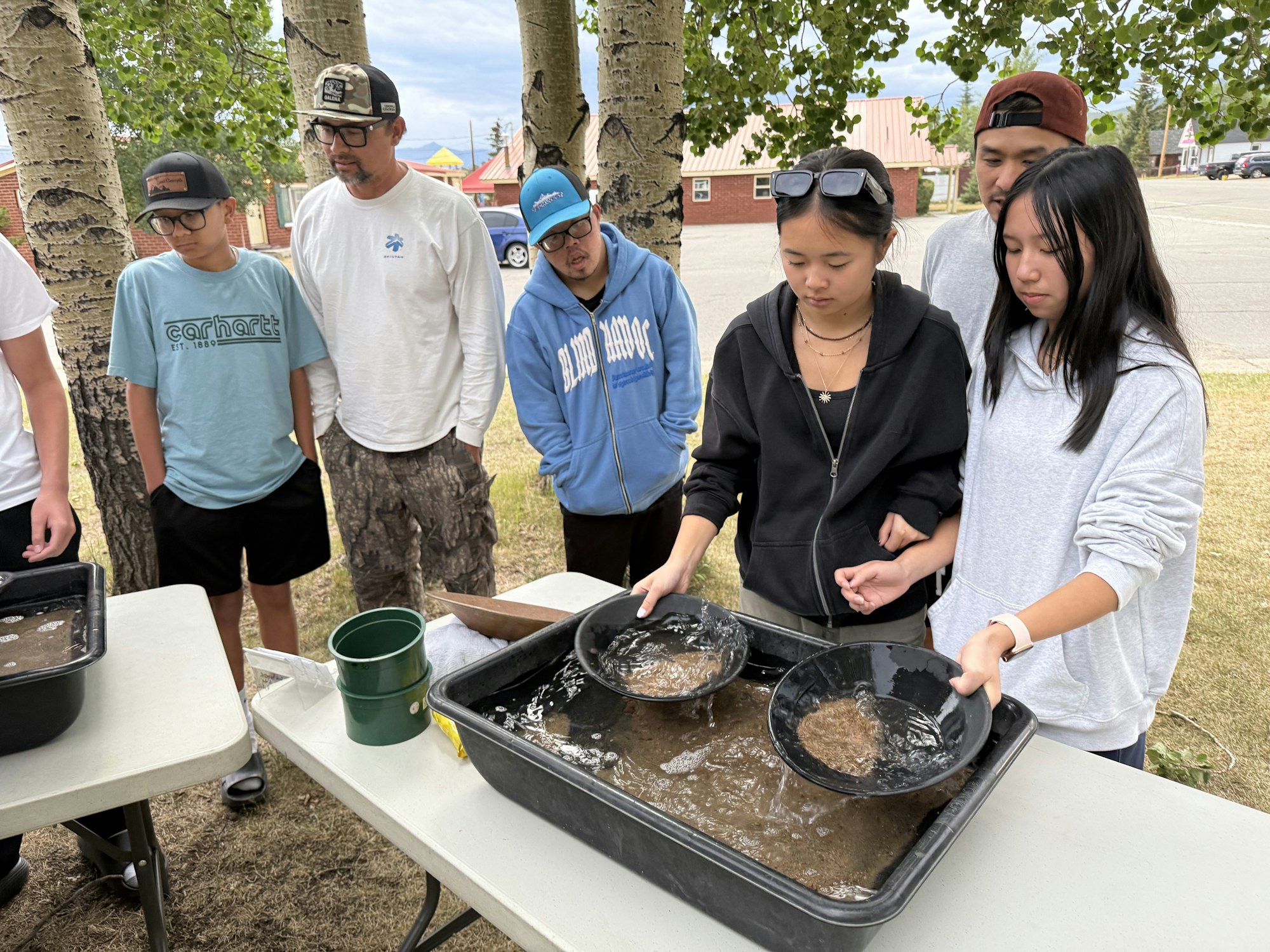 A group of people is panning for treasures in water at a table outdoors, with a few watching attentively.