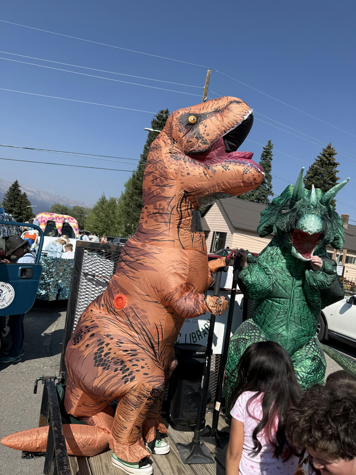 Two people in dinosaur costumes (a T-Rex and a green dinosaur) are seen at an outdoor event, with children nearby.