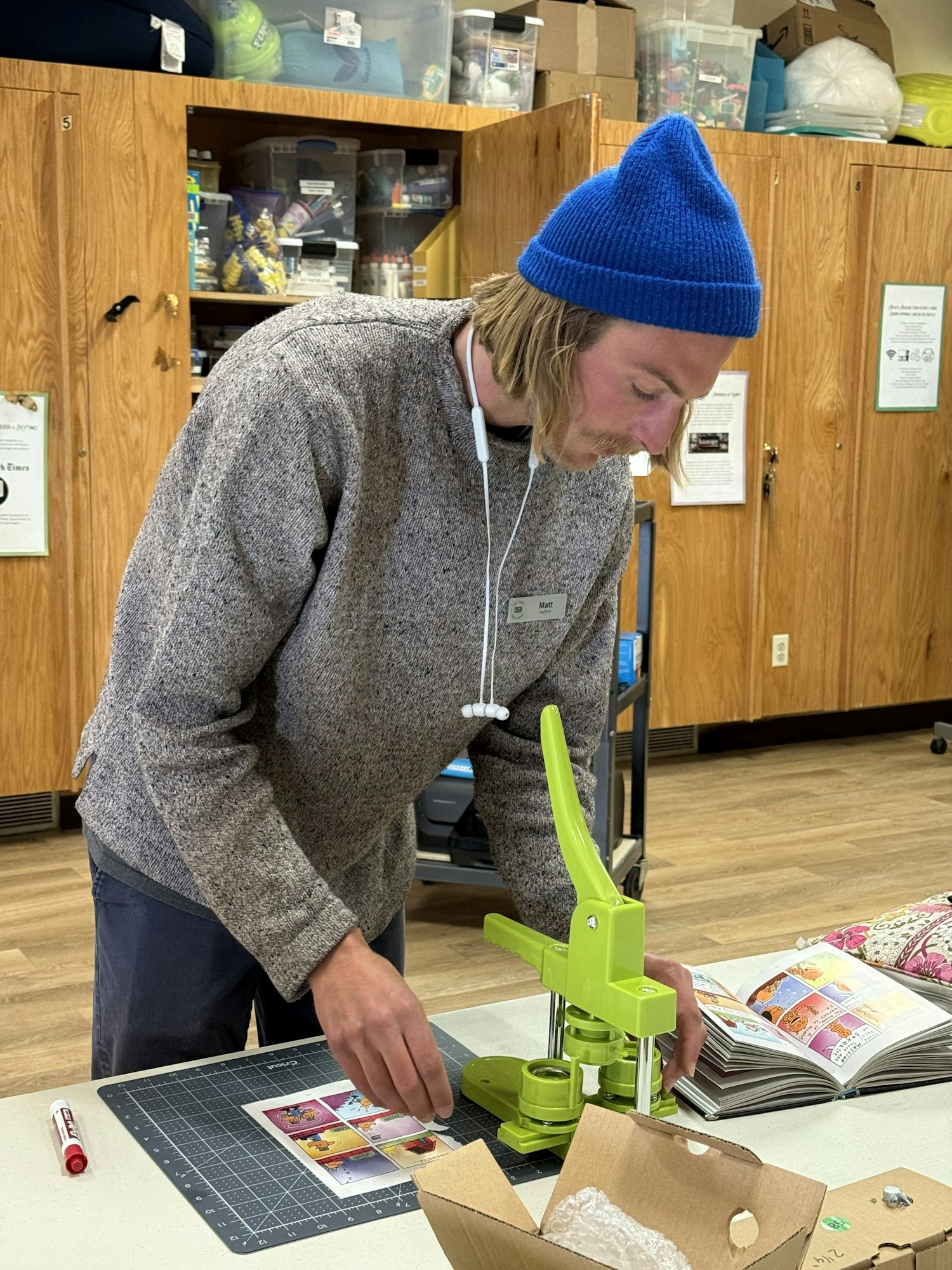 A person with a blue beanie works with a green press machine, arranging images on a table in a crafting space.
