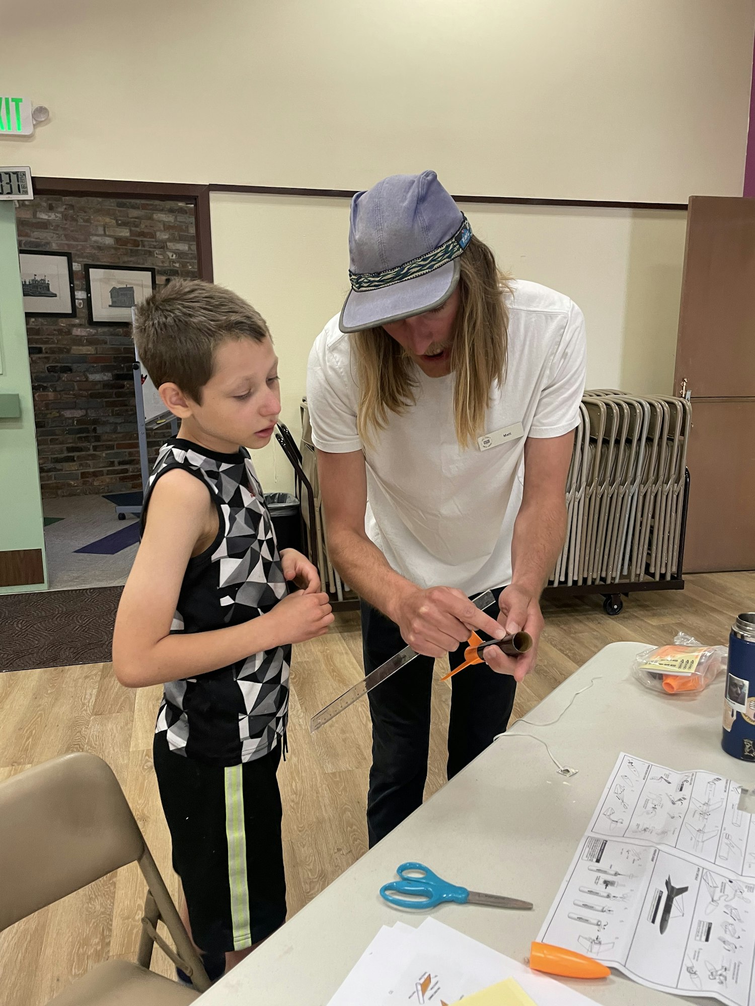 A boy and a man are working together on a project involving tools and materials at a table in a community space.