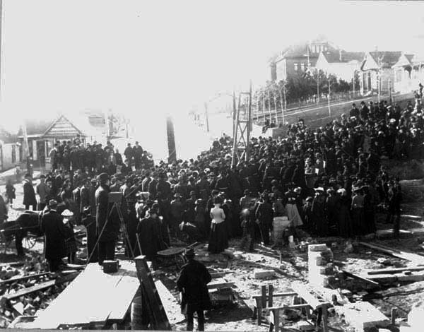 A crowd gathered at a construction site, possibly after an incident, with workers and onlookers in early 20th-century attire.
