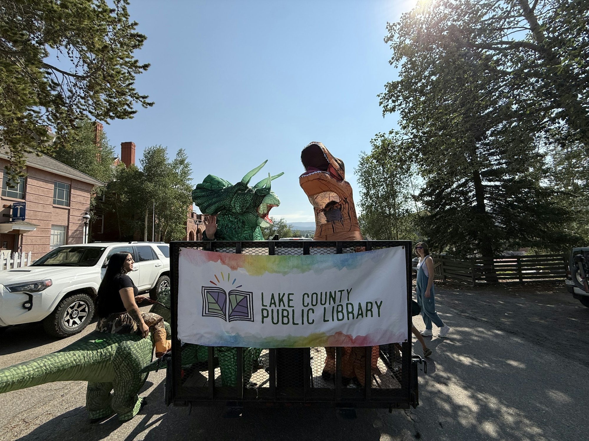 A trailer features people in dinosaur costumes promoting the Lake County Public Library on a sunny day.