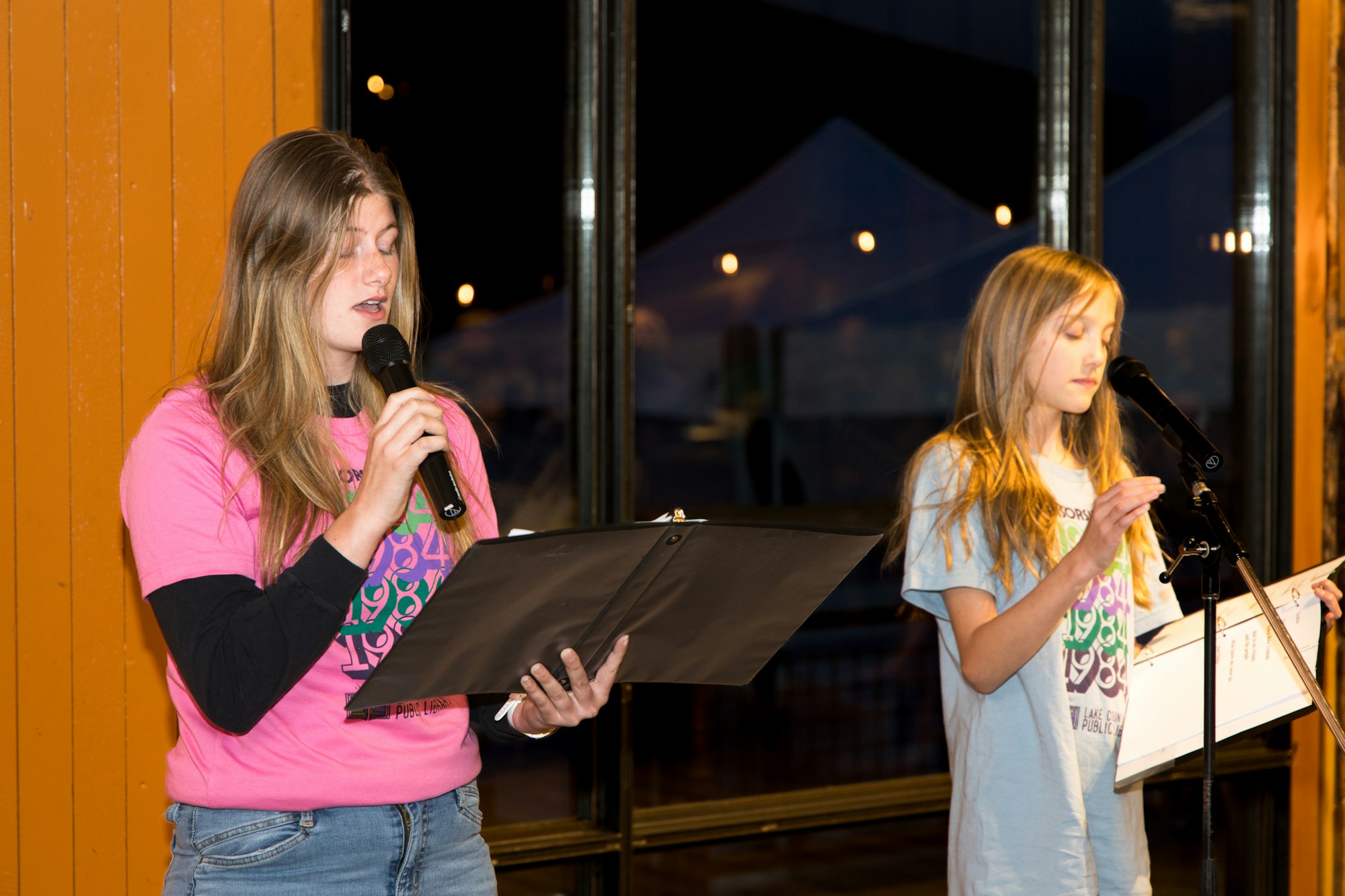 Two people with microphones reading from scripts, standing indoors.