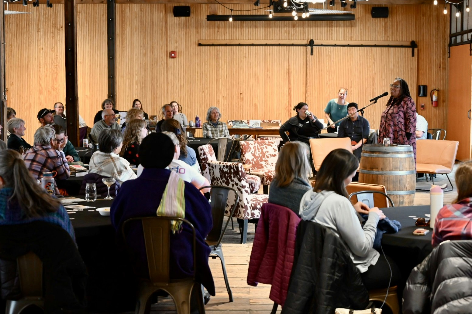A group of people is seated in a venue, with one person speaking at a microphone while others listen and engage.