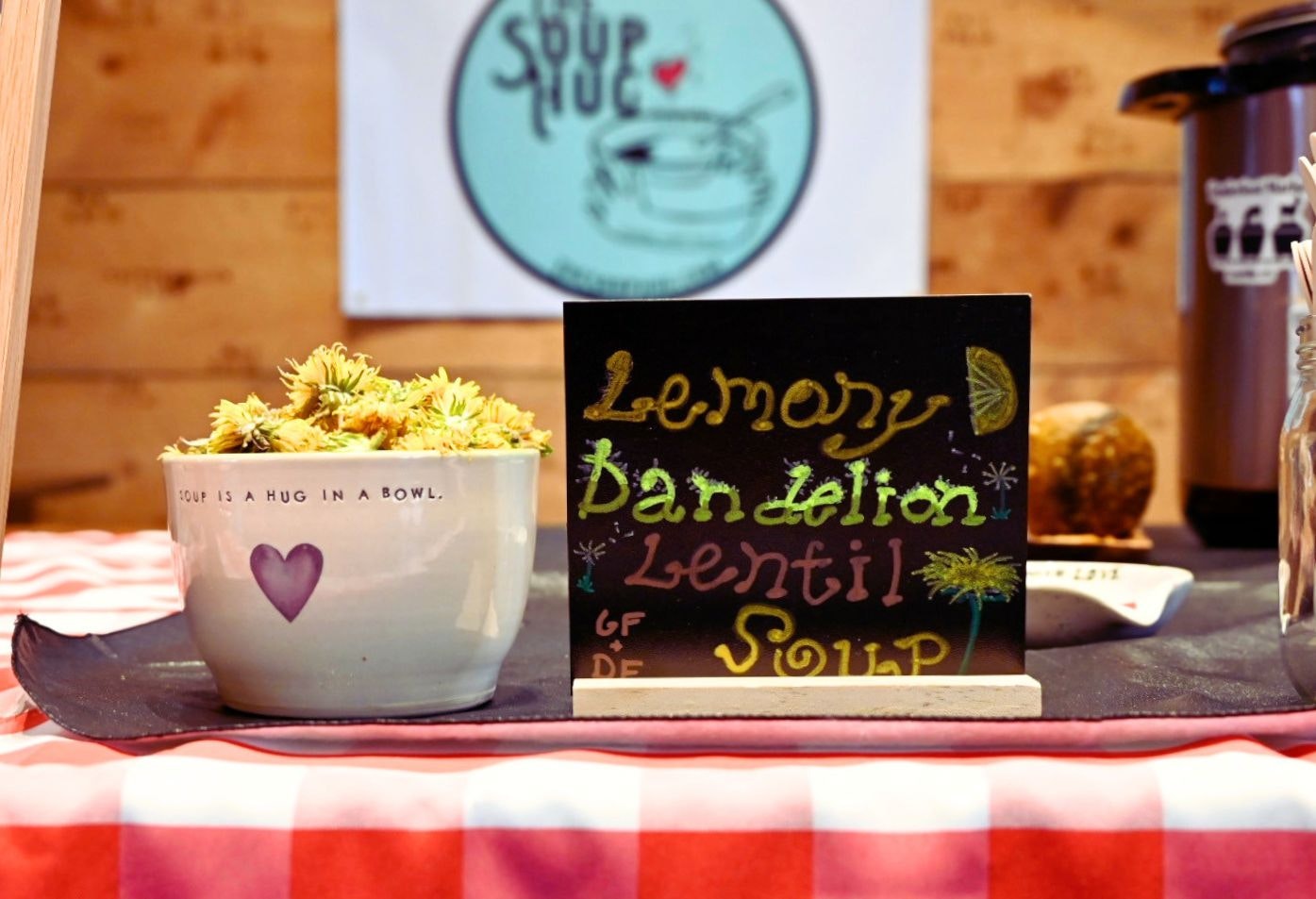 A bowl with yellow flowers and a sign for "Lemony Dandelion Lentil Soup" at a cozy soup stand. "Soup is a hug in a bowl."
