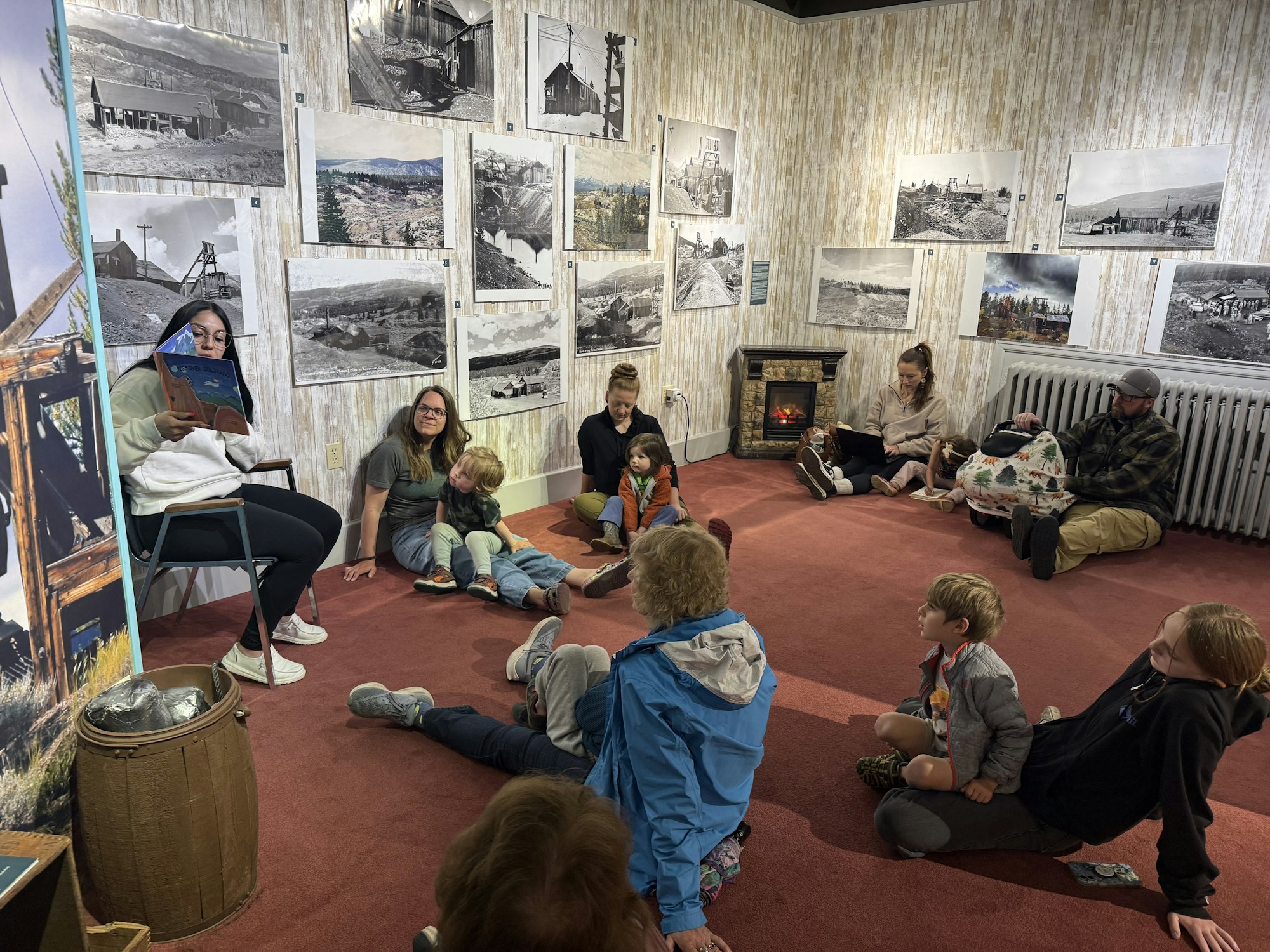 A group of children and adults sit in a cozy room while someone reads a story, surrounded by historical photos.