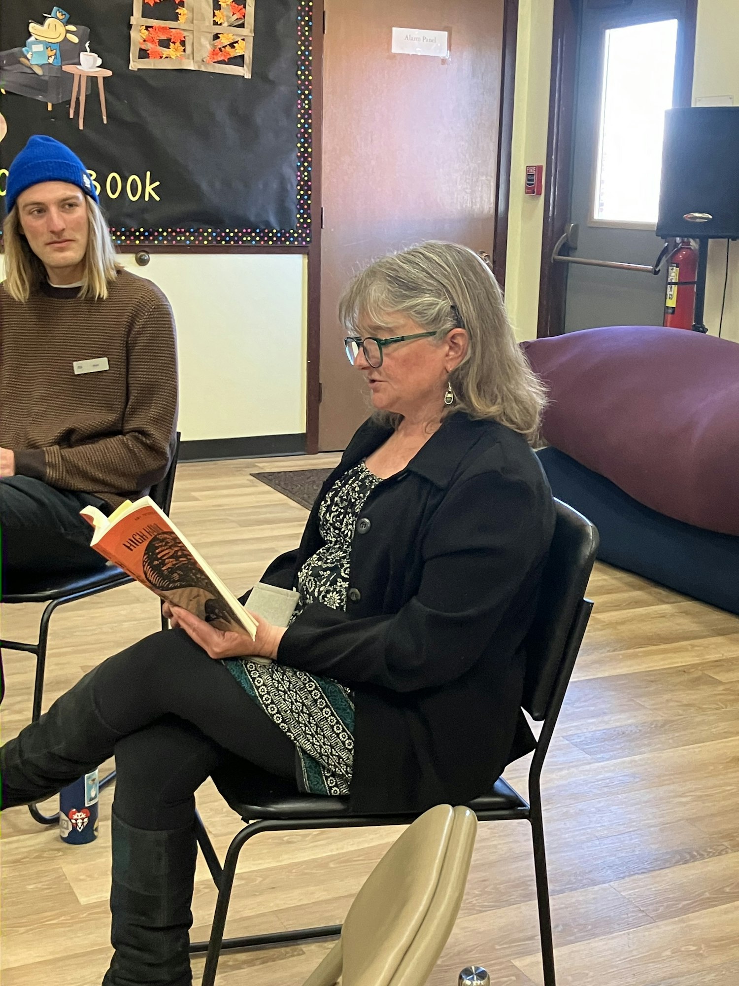 A woman reads a book while a man listens, in a cozy setting with a colorful backdrop and casual furniture.