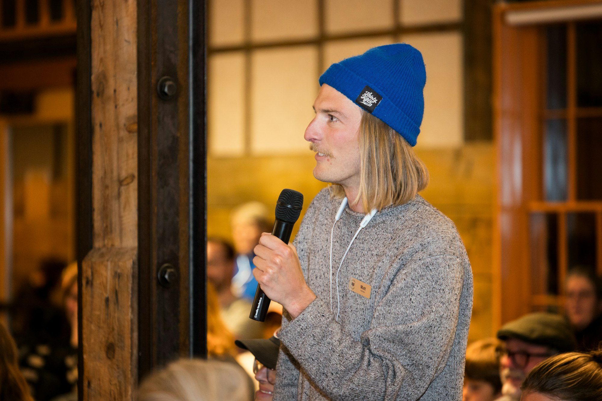 A person with long hair and a blue beanie speaks into a microphone, addressing an audience in a cozy indoor setting.