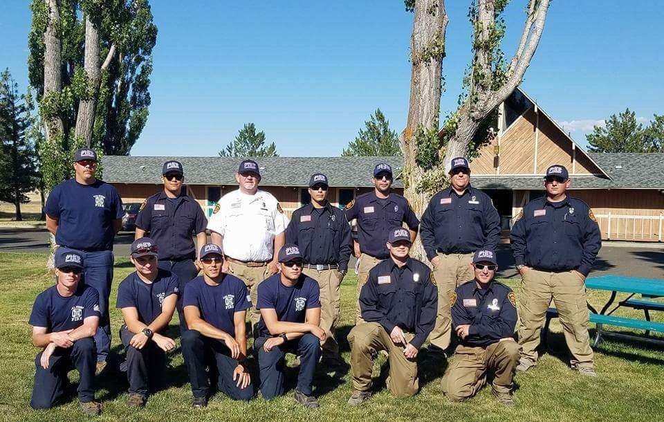 A group of firefighters and personnel, posing outdoors in uniforms, with trees and a building in the background.