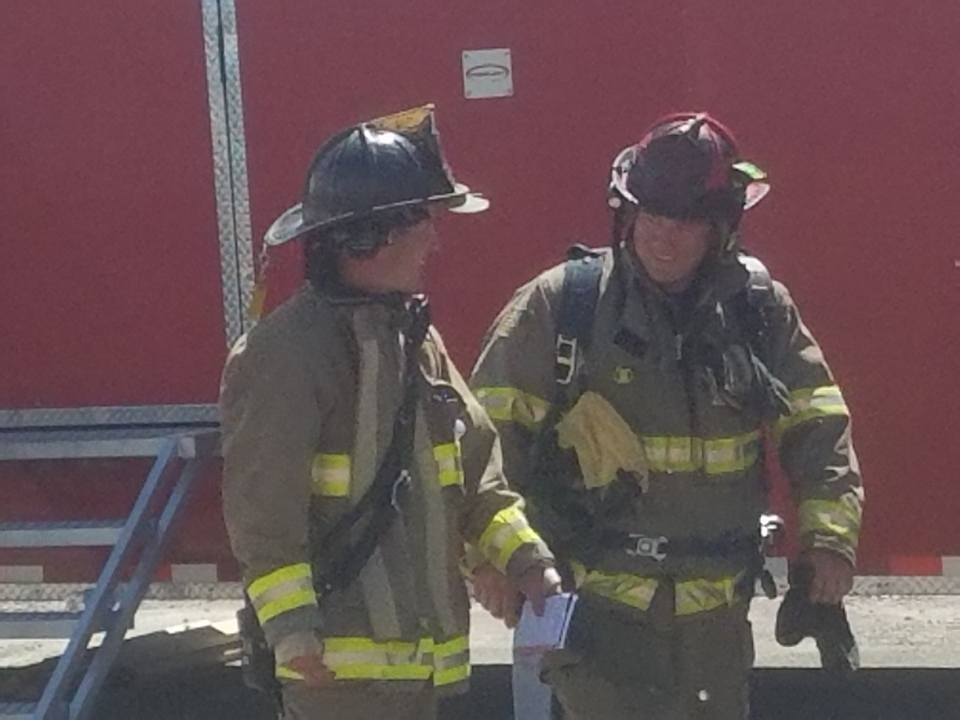 Two firefighters in gear talking in front of a red fire truck.