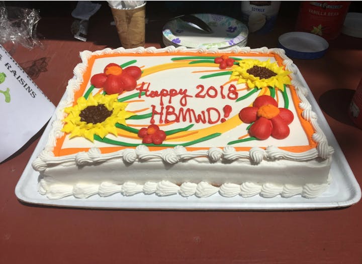 A colorful cake decorated with flowers, wishing "Happy 2018 HBMWD!" on top, placed on a table.