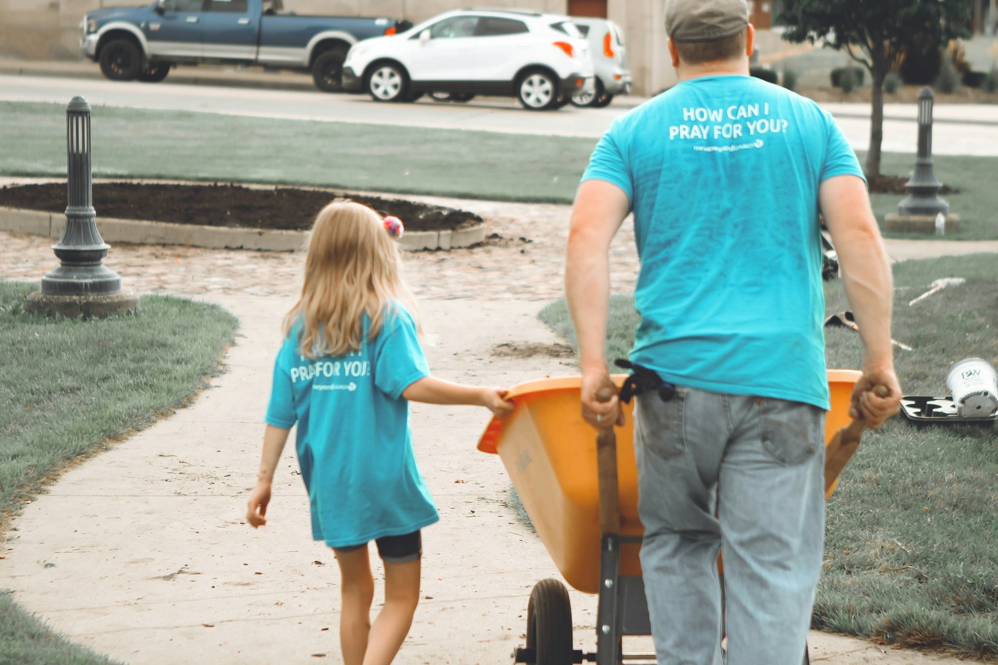 A child and an adult wearing matching blue shirts walk together, with the adult pulling a wheelbarrow in a grassy area.