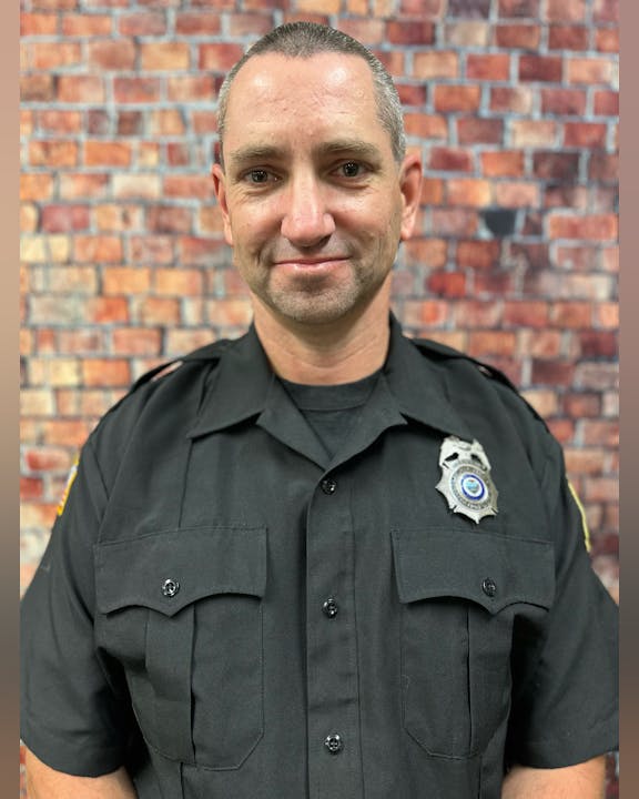 A person in a police uniform with a badge stands in front of a brick wall background.