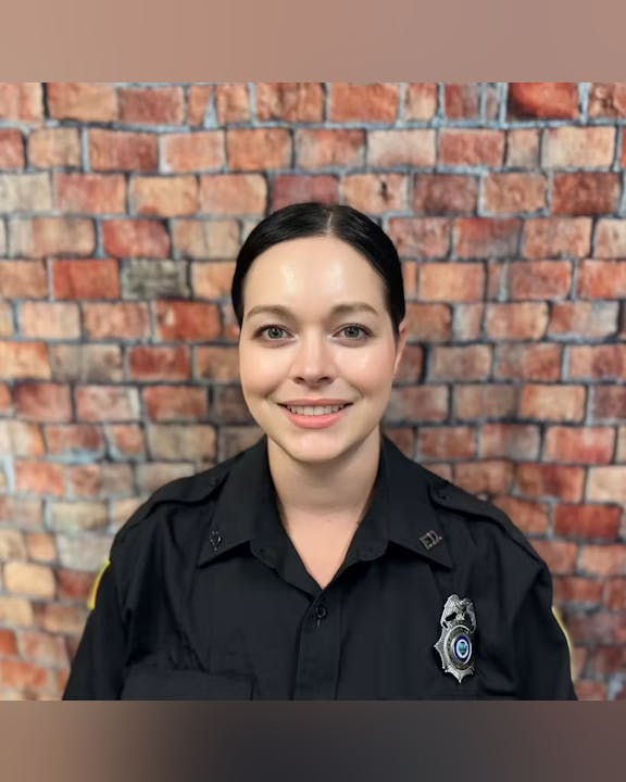 A person in a black uniform with a badge stands in front of a brick wall.