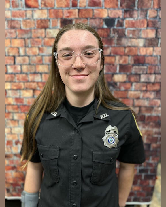A person smiling in police-like uniform with a badge, standing before a brick wall.