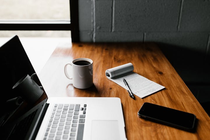 A wooden desk with a laptop, coffee mug, notepad, pen, and phone.