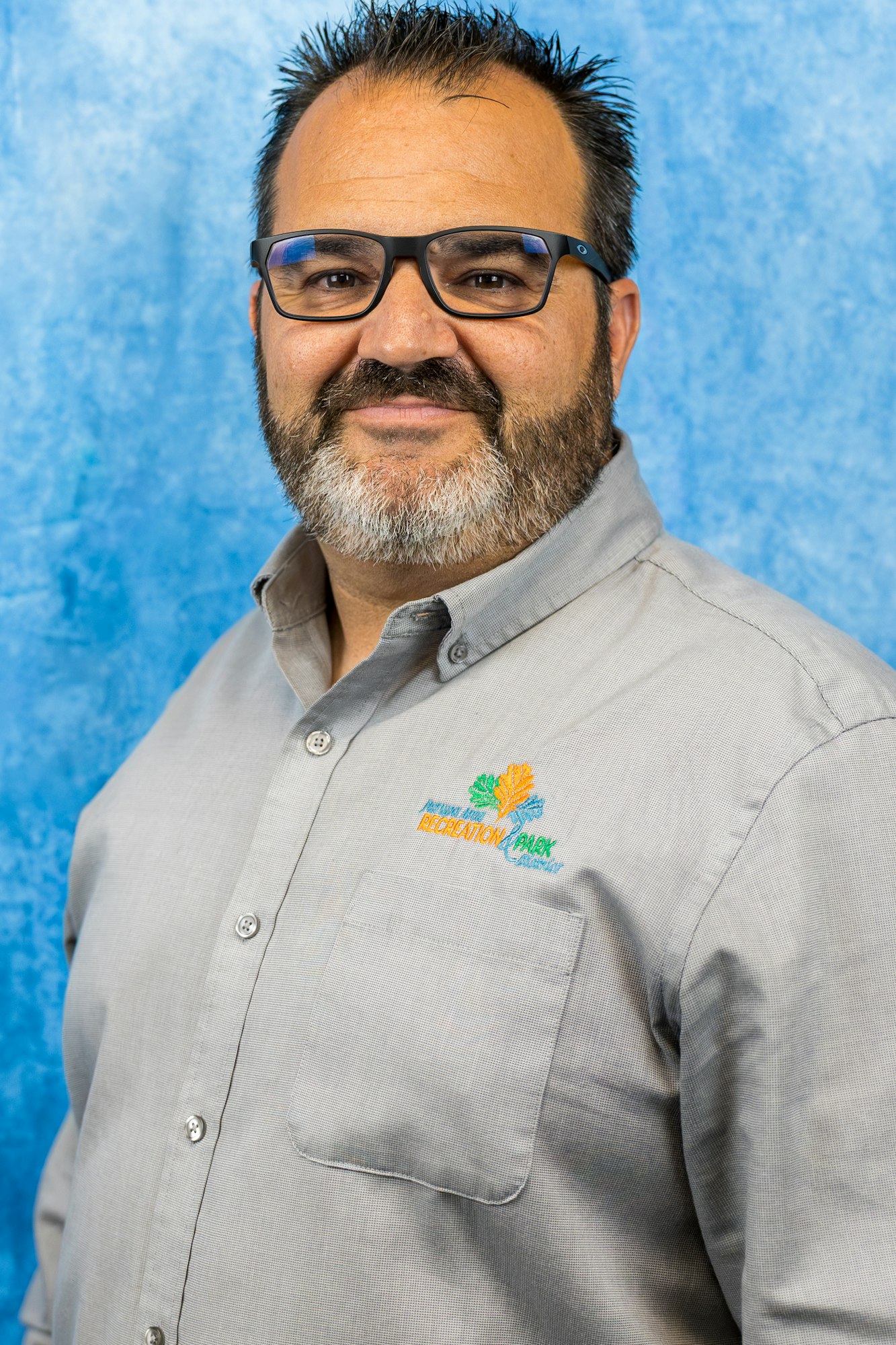 The image features a man with a beard and glasses, wearing a gray shirt with a colorful logo on it, set against a blue background.