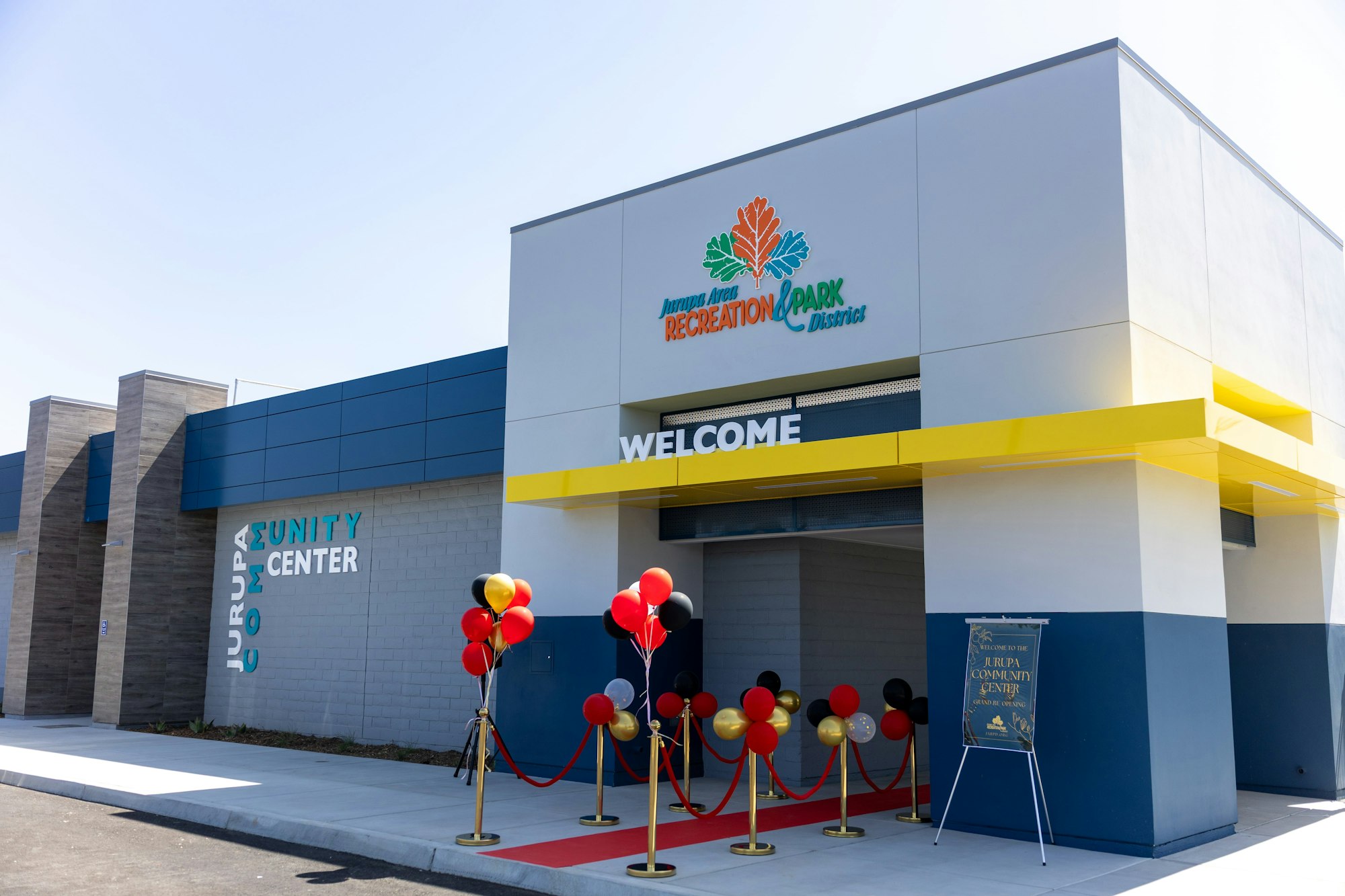 Modern community center entrance with "WELCOME" sign, red carpet, and red, black, and gold balloons.