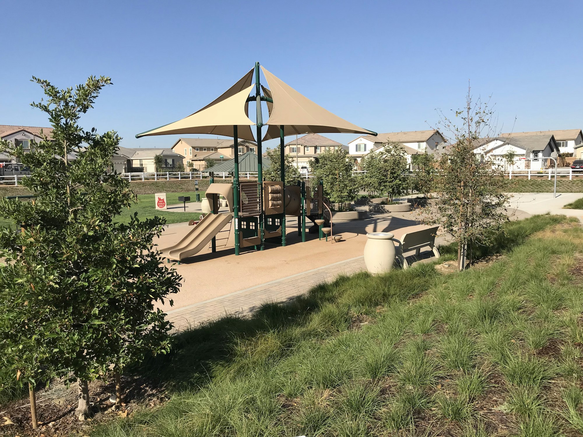 A sunny park scene featuring a playground with a slide, trees, grass, and nearby homes, perfect for outdoor play and relaxation.