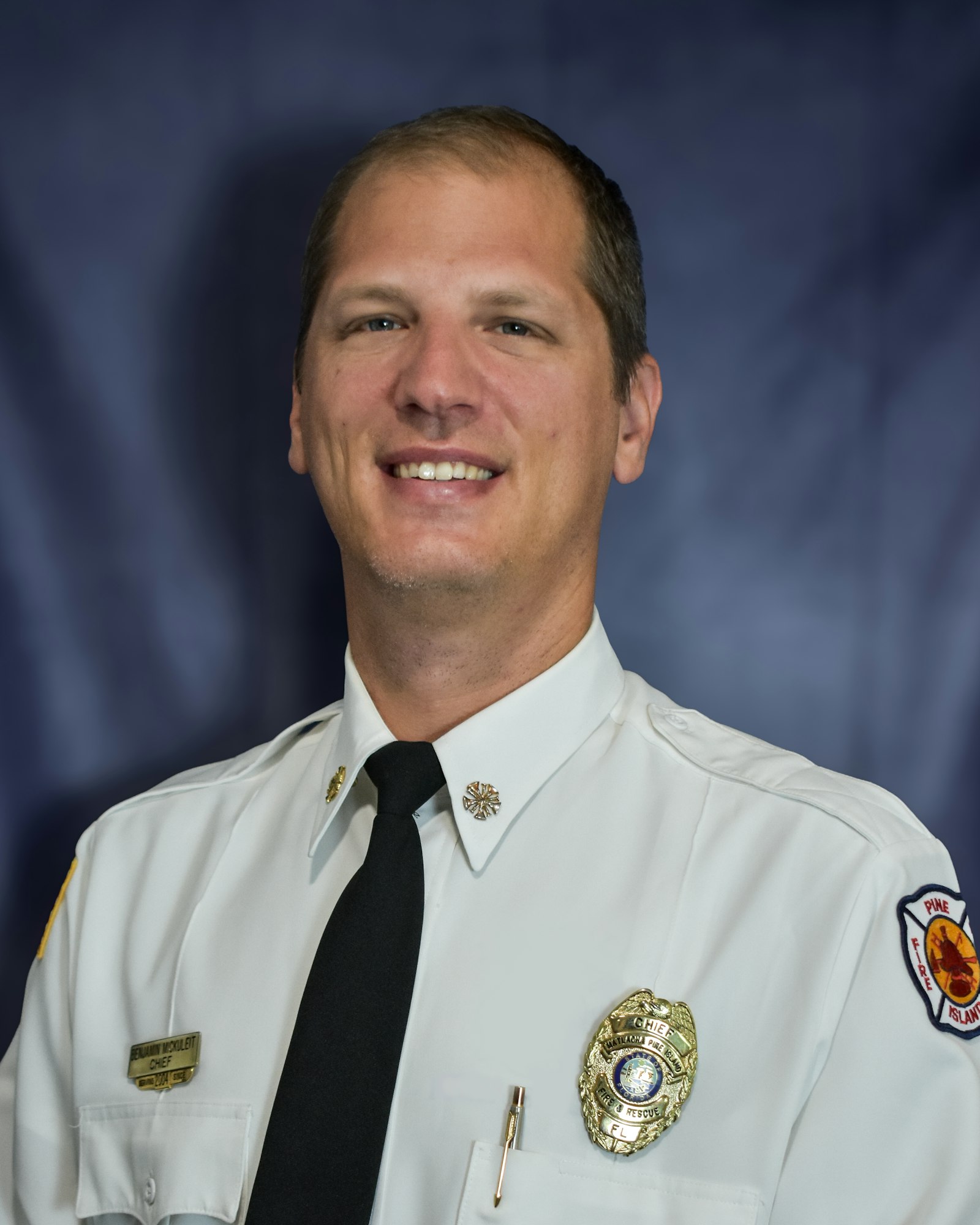 A man in a white uniform with badges and a tie, standing in front of a blue background.