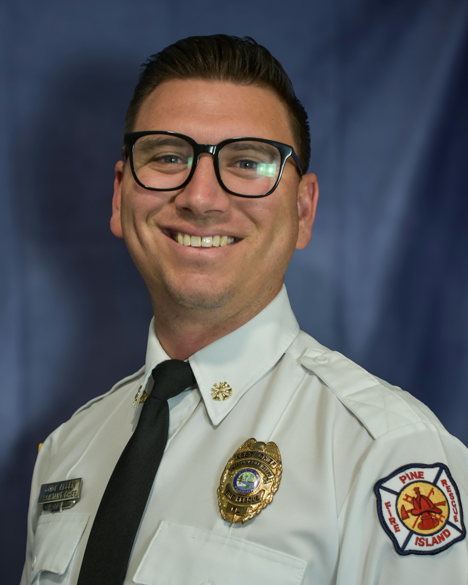 A smiling person in a fire department uniform, wearing glasses, with a badge and embroidered patch on the shirt.