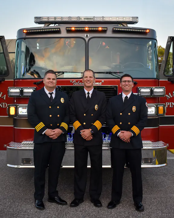 Three uniformed fire service personnel stand in front of a fire truck, smiling and posing for the photo.