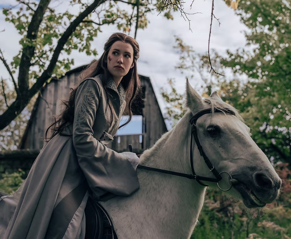 A person in medieval attire riding a white horse, set in a wooded area with an old wooden barn in the background.