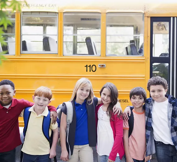 A group of six children standing in front of a yellow school bus, smiling with backpacks on.