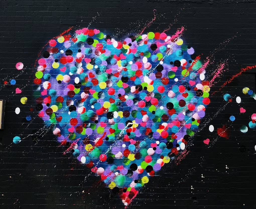 A colorful heart-shaped graffiti made of multicolored dots on a black brick wall.