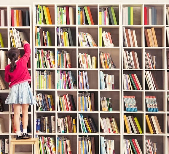 A girl standing on a stool reaches for a book on a tall, filled bookshelf.