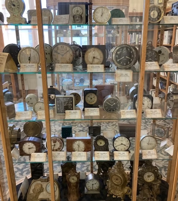 A display case filled with various vintage clocks, each with a descriptive label.