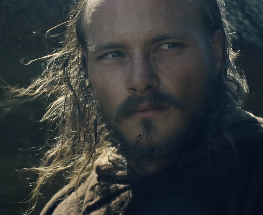 A bearded person with long hair looks off-camera, wearing a dark, rugged outfit.