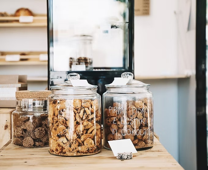 Jars filled with cookies on a wooden counter, some with labels, in a bakery or coffee shop setting.